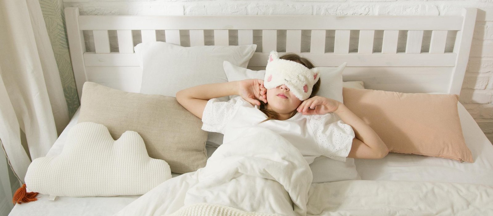 A young girl resting in bed with a cute sleep mask surrounded by cozy pillows.